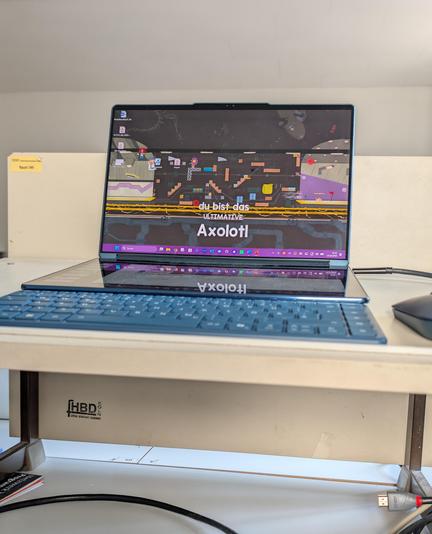 Foto meines Laptops auf einem Pult. Der Bildschirm hat als Hintergrund gerade einen Screenshot aus dem Spiel "Ultimate Chicken Horse" und daraus steht "You are the ultimate Axolotl".