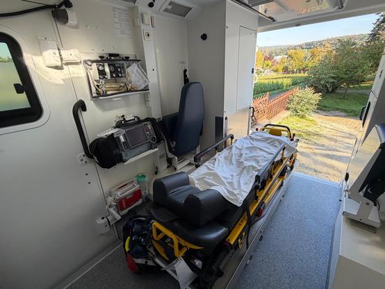 Blick von Innen in den hellen Behandlungsraum des Rettungswagens mit einer Rettungstrage im Vordergrund, medizinischen Geräten an der Seitenwand, einem integrierten Sitz und dem Blick durch die geöffnete Hecktür ins Freie.